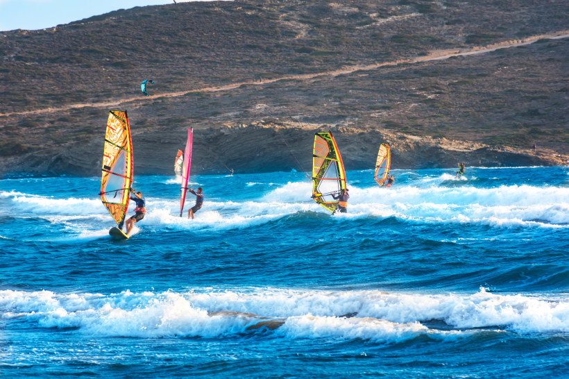 Das Bild zeigt eine lebhafte Szene mit mehreren Windsurfern auf einem aufgewühlten Meer mit kleinen bis mittelhohen Wellen.   Die Sportler nutzen farbenfrohe Segel in Gelb, Pink und Orange und bewegen sich mit hoher Geschwindigkeit über das Wasser.   Im H
