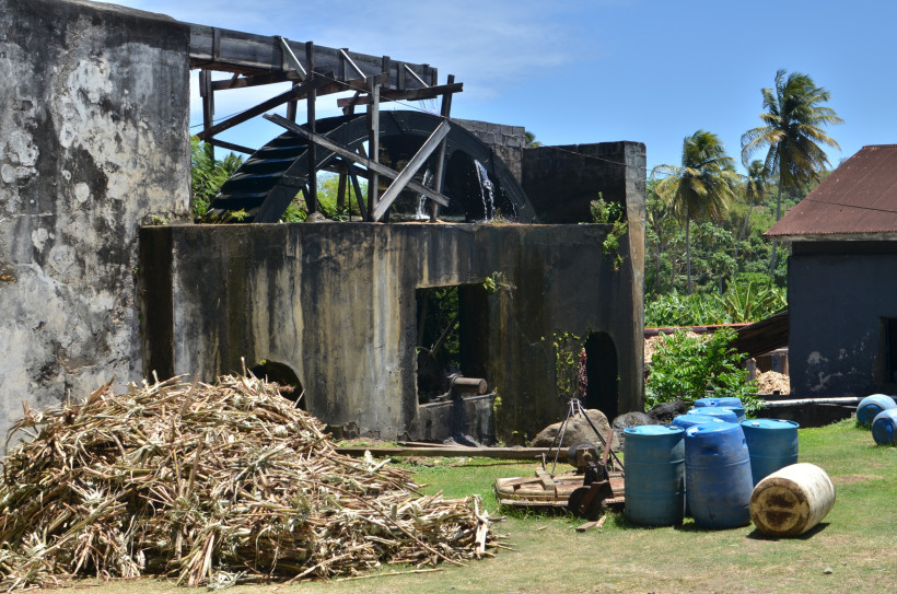 Curaçao - Liqueur Distillery Außenansicht einer alten Zuckermühle mit großem hölzernem Wasserrad. Im Vordergrund liegen Zuckerrohrreste auf dem Boden, daneben stehen blaue Plastikfässer und einige Werkzeuge. Im Hintergrund Palmen und tropische Vegetation unter blauem Himmel.