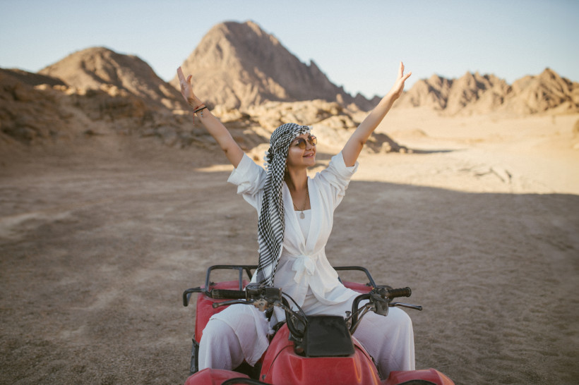 Ägypten - Makadi Bay Eine lächelnde Frau sitzt auf einem roten Quad inmitten einer Wüstenlandschaft mit Felsen und Bergen im Hintergrund. Sie trägt weiße Kleidung, eine Sonnenbrille und ein schwarz-weißes Tuch um den Kopf. Die Arme hat sie weit nach oben gestreckt, als Ausdru