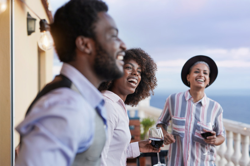 Südafrika Drei Personen stehen auf einer Terrasse mit Meerblick, lachen und unterhalten sich. Zwei von ihnen halten Gläser mit Rotwein in der Hand. Die Stimmung ist gelöst und freundlich
