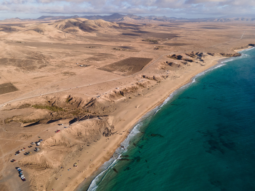 Luftaufnahme von Piedra Playa mit Sandstrand, Felsküste und türkisblauem Meer vor einer trockenen Hügellandschaft.
