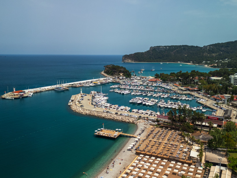 Panoramablick auf die Kemer Marina mit Hafenbecken, zahlreichen Booten, Strandliegen und Bergen im Hintergrund