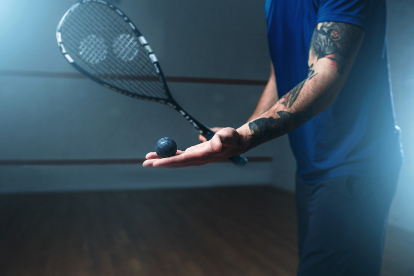 Ägypten - Marsa Alam Nahaufnahme eines Squashspielers mit tätowiertem Arm, der einen Squashball in der geöffneten Hand hält und einen Schläger in der anderen. Der Hintergrund ist leicht verschwommen.