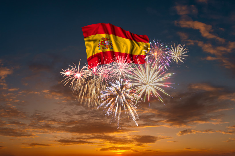 Ibiza Spanische Flagge vor nächtlichem Himmel, umgeben von farbenfrohem Feuerwerk. Im Hintergrund geht die Sonne unter, der Himmel ist in orange und blau gefärbt