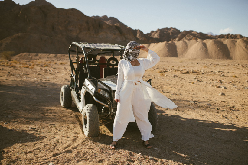 Ägypten - Hurghada Eine Frau steht in einem weißen Outfit und mit Tuch um den Kopf neben einem Buggy in einer weiten, sonnigen Wüstenlandschaft. Im Hintergrund sind felsige Hügel zu sehen.