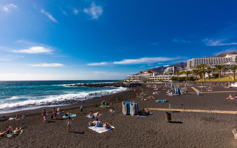 Teneriffa - Playa de la Arena Playa de la Arena in Puerto de Santiago, Teneriffa: schwarzer Vulkansand mit Badegästen, Atlantik-Wellen, Promenade mit Hotels und Palmen, Berge unter blauem Himmel.