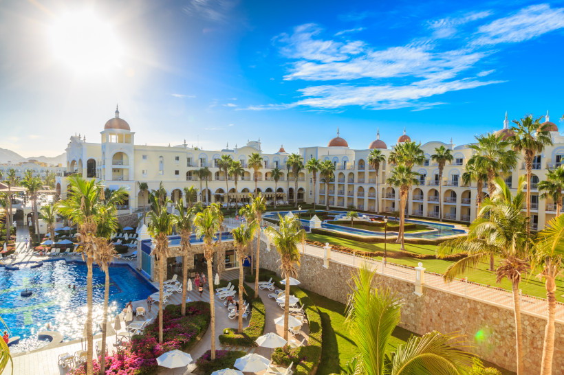 Ägypten - Hurghada Sonnenbeschienenes, elegantes Hotelresort mit mehreren Etagen, weißer Fassade und Kuppeln. Im Vordergrund befinden sich Palmen, ein Poolbereich mit Liegestühlen sowie gepflegte Gartenanlagen. Der Himmel ist blau mit wenigen Wolken