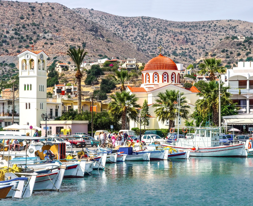 Hafen von Elounda mit Fischerbooten im Vordergrund, Kirche mit roter Kuppel und Glockenturm vor Hügellandschaft