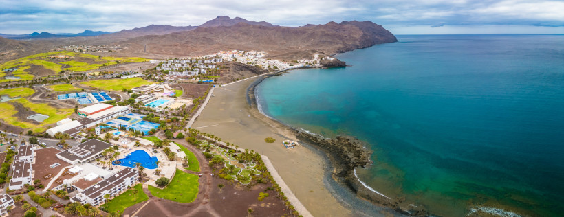 Panoramablick auf die Bucht von Las Playitas mit Sportanlagen und Bergen Die Aufnahme zeigt Las Playitas aus der Vogelperspektive: die geschützte Bucht mit dunklem Sand, das große Sport- und Hotelresort im Vordergrund und die dramatischen Bergketten im Hintergrund. Ein typisch ostkanarischer Mix aus Ruhe, Weite und sportlicher