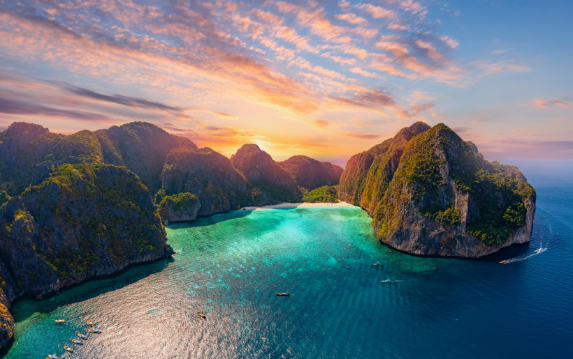Bucht mit türkisfarbenem Wasser, weißem Sandstrand und von steilen, grünen Felsen umgeben. Der Sonnenuntergang färbt den Himmel in orange-rosa Tönen. Kleine Boote liegen im Wasser.