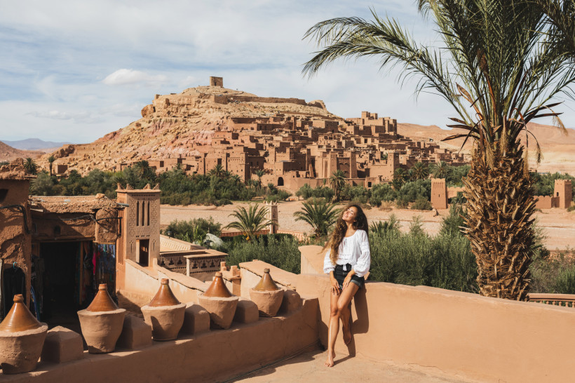 Marokko Frau lehnt entspannt an einer Mauer mit Blick auf die historische Lehmstadt Aït-Ben-Haddou in Marokko. Umgeben von Palmen, traditionellen Gebäuden und rötlich-gelben Felsen in der Wüstenlandschaft.