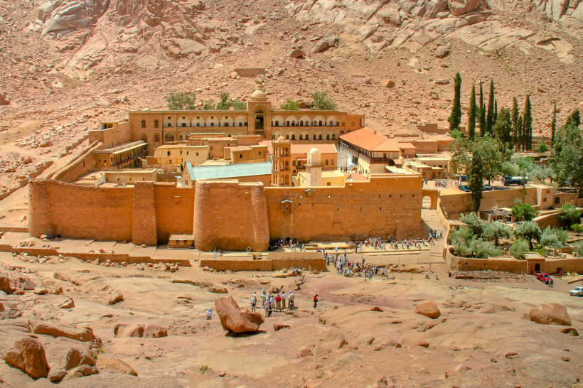 Ägypten - Sharm el Sheikh Das Katharinenkloster am Fuße des Berges Sinai, umgeben von rötlichen Felsen und karger Landschaft. Die massive Klosteranlage mit ihren hohen Mauern und Gebäuden in warmen Gelbtönen ist von zahlreichen Besuchern umgeben. Im Hintergrund ragen Zypressen und