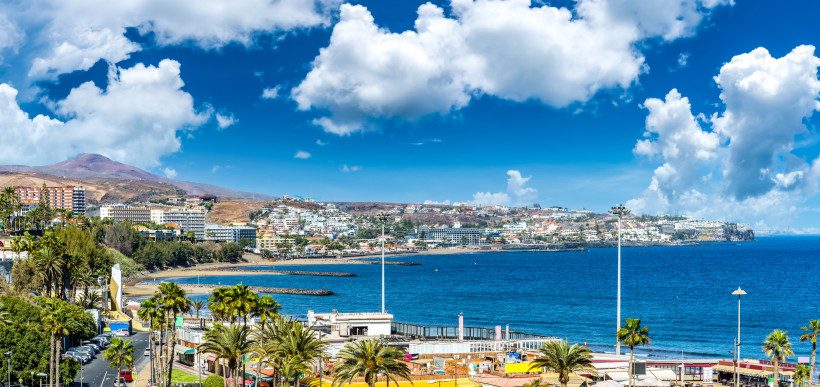 Blick auf San Agustín auf Gran Canaria mit Hotels, Apartmentanlagen und grünen Palmen entlang der Küste vor tiefblauem Atlantik und malerischem Wolkenhimmel.