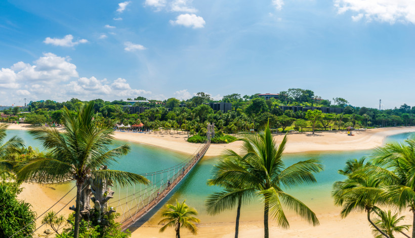 Singapur Panoramablick auf einen tropischen Strand auf Sentosa Island in Singapur. In der Mitte des Bildes verläuft eine schmale Hängebrücke über eine türkisfarbene Lagune, die von goldgelbem Sand und üppigen Palmen gesäumt ist. Im Hintergrund sieht man dichten, g