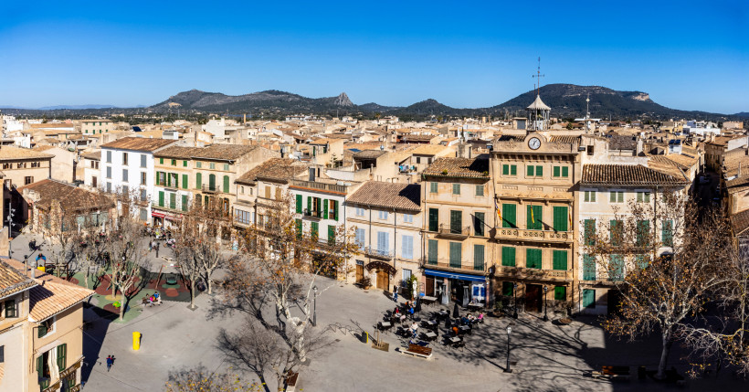 Plaça d’Espanya mit historischen Fassaden, Uhrenturm und Platzfläche mit Bäumen und Sitzgelegenheiten