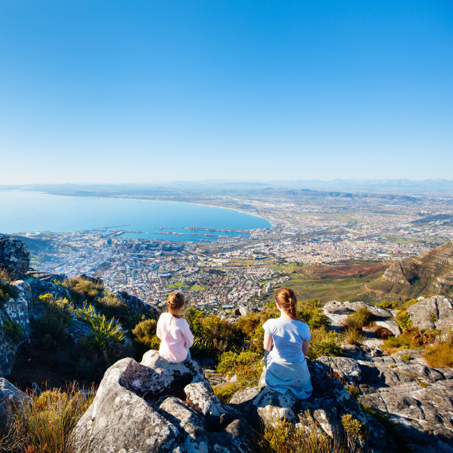 Südafrika Zwei Personen sitzen auf einem Felsen hoch oben auf einem Berg und blicken auf die südafrikanische Stadt Kapstadt. Die Stadt liegt direkt am Meer, der Blick reicht über die Bucht bis zum Horizont. Es ist ein klarer, sonniger Tag, der Himmel ist wolkenlos
