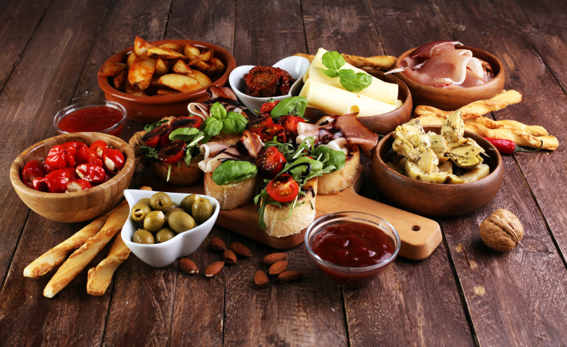 Tapas-Auswahl auf Holztisch mit Brot, Schinken, Käse, Artischocken, Oliven, Bratkartoffeln und Dips