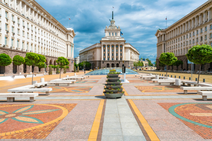 Blick auf das Largo in Sofia, ein monumentaler Platz mit drei großen Regierungsgebäuden im sozialistischen Klassizismus. In der Mitte ragt das Parlamentsgebäude mit Flagge und Turm auf. Der Platz ist mit farbigem Mosaikpflaster gestaltet, gesäumt von klei