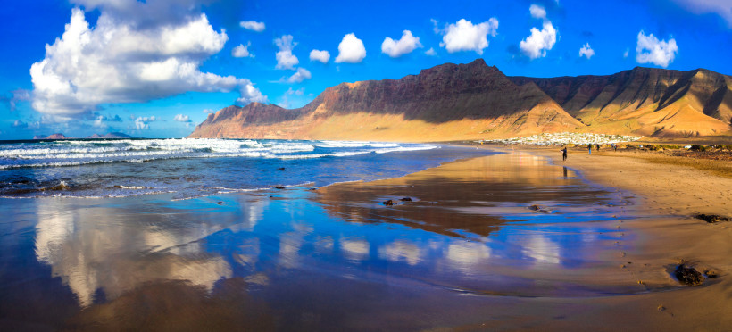 Playa de Famara: Lanzarotes beeindruckender Naturstrand vor majestätischer Klippenkulisse Blick auf den weiten Playa de Famara mit reflektierenden Wellen und den imposanten Famara-Klippen auf Lanzarote