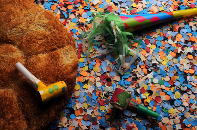Buntes Konfetti bedeckt den Boden, daneben liegen Partytröten und ein großes, rundes Gebäck. Die Szene wirkt festlich und erinnert an Karneval oder eine Feier