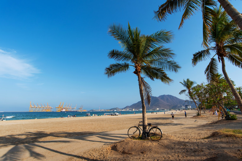 Fujairah, UAE Tropischer Strand mit Palmen, einem angelehnten Fahrrad, Menschen am Ufer und Hafenkränen im Hintergrund