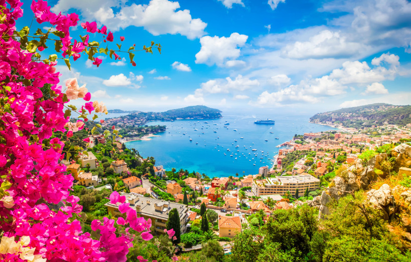 Frankreich Panorama einer mediterranen Bucht mit pastellfarbenen Häusern, Bougainvillea im Vordergrund und zahlreichen Booten bis zum Horizont.