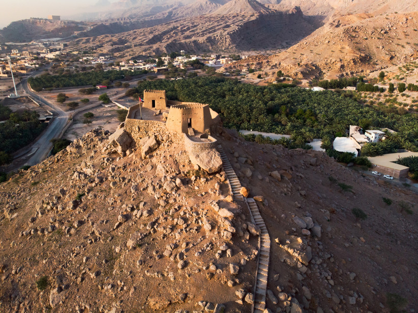 Luftaufnahme des Dhaya Fort in Ras Al Khaimah – historische Festung auf einem Hügel mit Palmenhainen und Gebirgspanorama.