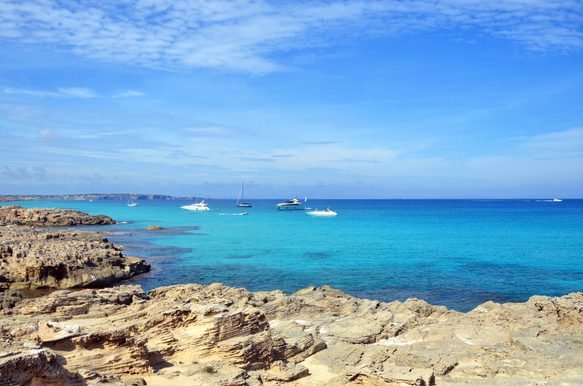Formentera - Es Caló de Sant Agustí Felsige Küste auf Formentera mit weitem, türkisblauem Meer; mehrere Yachten und Segelboote liegen vor Anker, klarer Horizont unter leicht bewölktem, blauem Himmel.