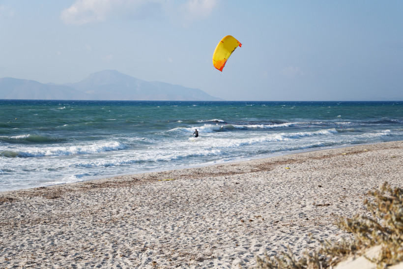Kos - Mastichari Mastichari Beach auf Kos: Kitesurfer mit gelb-rotem Schirm vor welligem, türkisblauem Meer und Bergkulisse.