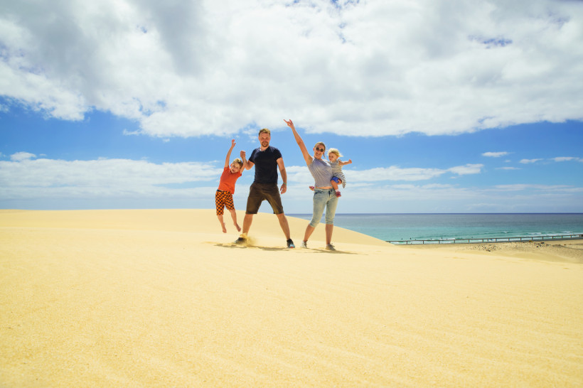 Fuerteventura Lachende Familie mit zwei Kindern steht auf einer goldgelben Sanddüne vor blauem Himmel und Meer. Ein Kind wird hochgehoben, das andere auf dem Arm getragen.