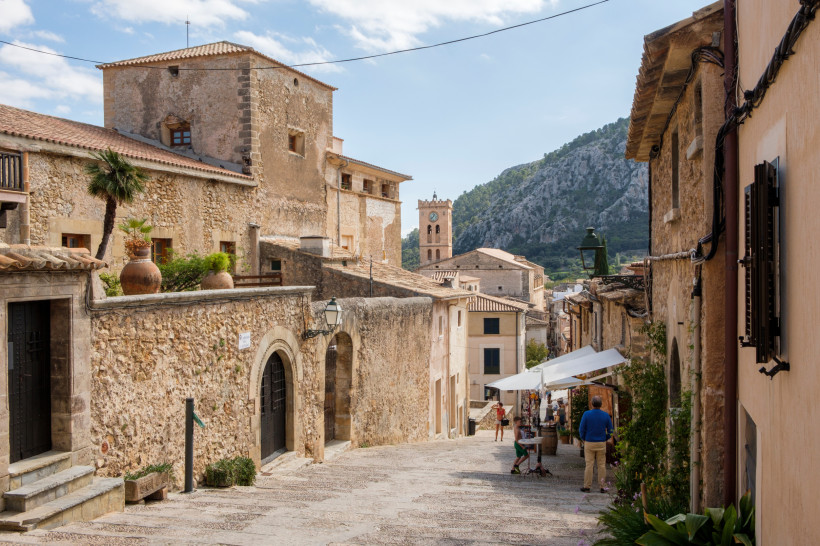 Gasse in der Altstadt von Pollença mit Natursteinhäusern, Marktständen und Blick auf einen Kirchturm vor Bergen