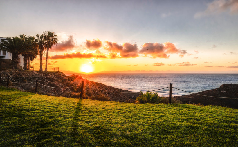 Fuerteventura Sonnenuntergang über dem Meer, gesehen von einer grünen Rasenfläche mit Palmen und Felsen im Vordergrund. Der Himmel ist orange-rosa gefärbt mit vereinzelten Wolken.