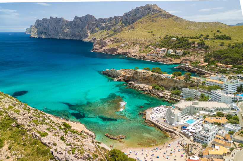 Blick über die Bucht von Cala Sant Vicenç mit türkisfarbenem Wasser, Sandstrand, Gebäuden am Ufer und felsigen Bergen im Hintergrund