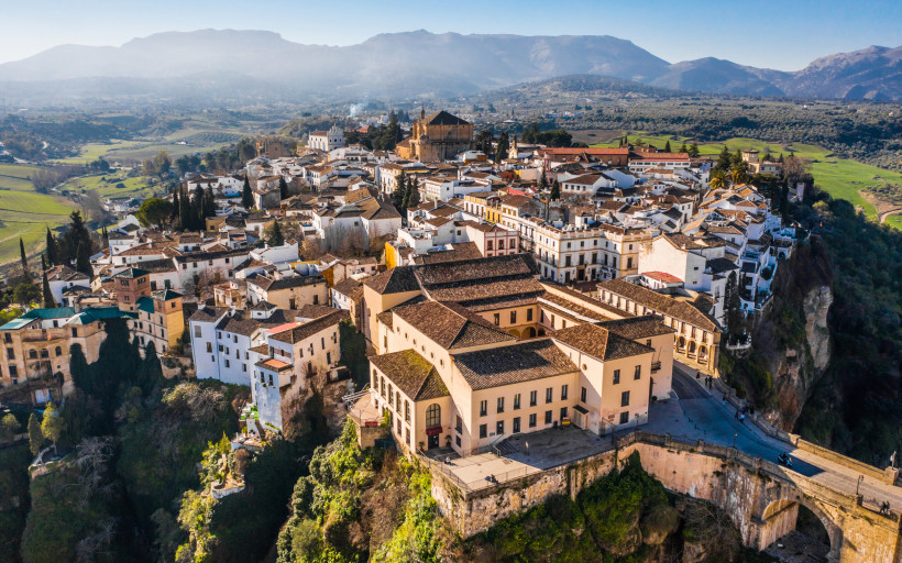 Luftaufnahme von Ronda in Andalusien, Spanien. Die weiße Altstadt liegt spektakulär auf einem Felsplateau, umgeben von grüner Landschaft und Bergen im Hintergrund.