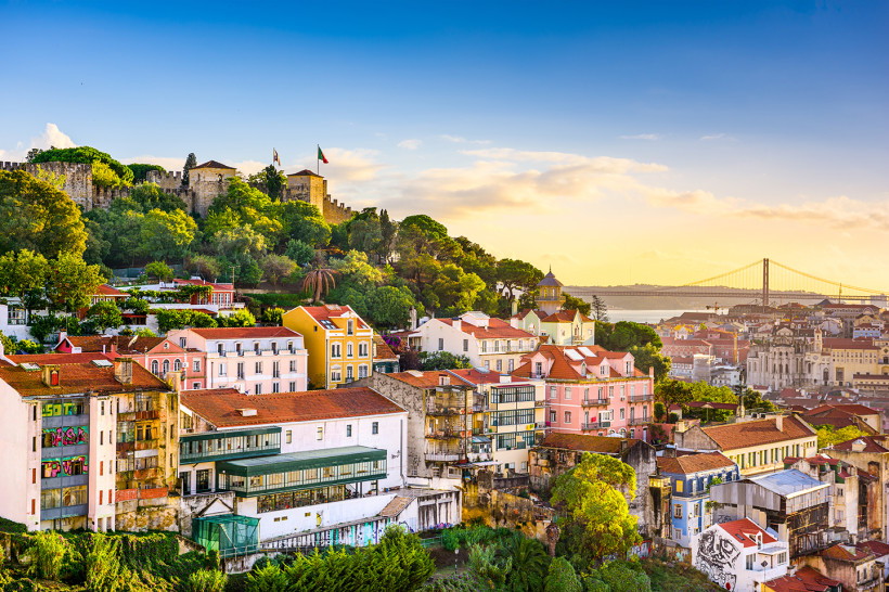 Lissabon Panoramablick auf Lissabon bei Sonnenuntergang. Im Vordergrund farbenfrohe Häuser mit roten Dächern, im Hintergrund das Castelo de São Jorge auf einem bewaldeten Hügel. Rechts im Bild die Hängebrücke Ponte 25 de Abril über dem Tejo-Fluss.