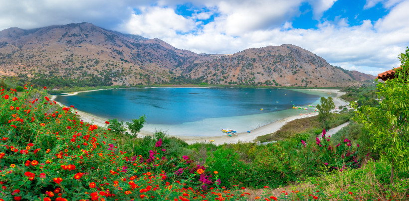 Panoramablick auf den Kournas-See mit sandigem Ufer, Bergen im Hintergrund und Blumen im Vordergrund