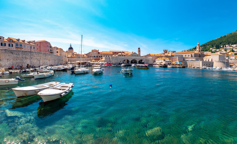 Blick auf den alten Hafen von Dubrovnik mit kleinen Booten und Yachten im klaren, türkisfarbenen Wasser. Dahinter historische Gebäude mit roten Dächern und Stadtmauer, umrahmt von Hügeln.