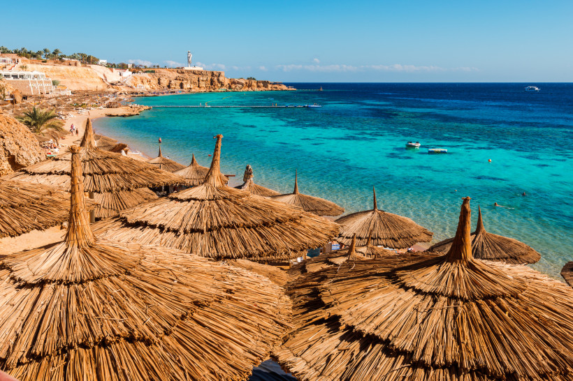 Ägypten Blick auf eine Bucht mit türkisfarbenem Wasser in Sharm el Sheikh. Im Vordergrund zahlreiche große Strohdach-Sonnenschirme, die sich terrassenförmig über den Hang erstrecken. Unten am Strand baden Menschen im Meer, während kleine Boote auf dem Wasser trei