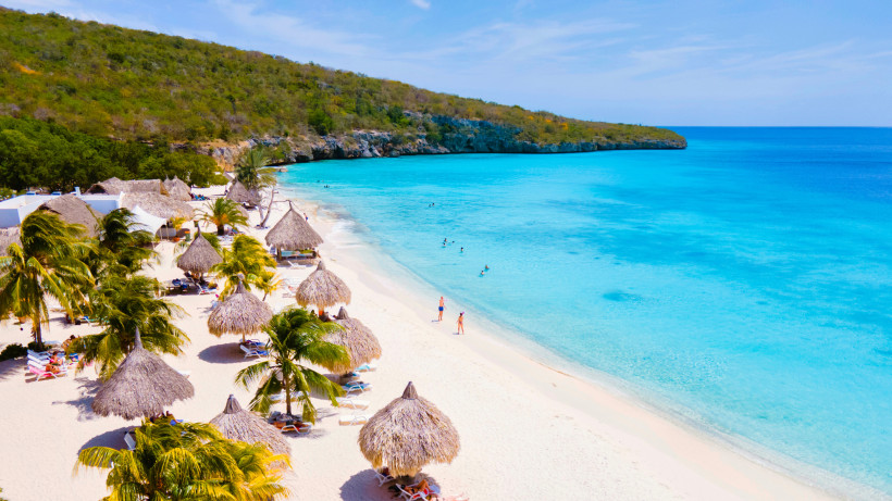 Strandbucht auf Curaçao mit weißem Sand, Palmen, Strohdach-Sonnenschirmen und türkisfarbenem Meer