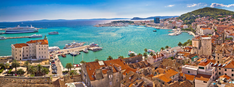 Panoramablick auf die Altstadt von Split in Kroatien mit roten Ziegeldächern, dem Hafen voller Yachten und Kreuzfahrtschiffen sowie türkisblauem Meer im Hintergrund. Entlang der Uferpromenade reihen sich Palmen und historische Gebäude.