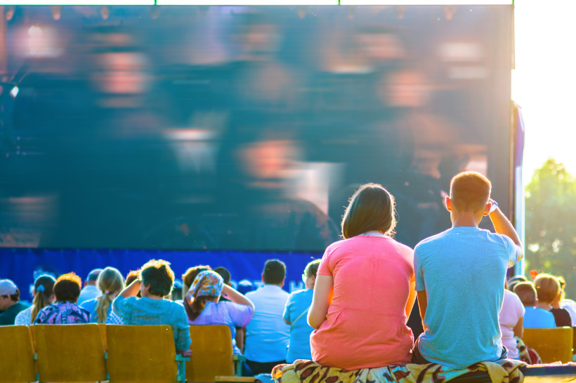 Mehrere Menschen sitzen auf Holzstühlen vor einer großen Freiluft-Leinwand bei Sonnenuntergang. Im Vordergrund ein junges Paar von hinten, er im blauen T-Shirt, sie im pinkfarbenen Kleid. Die Leinwand zeigt einen unscharfen Film in dunklen Farben. Sonnenl