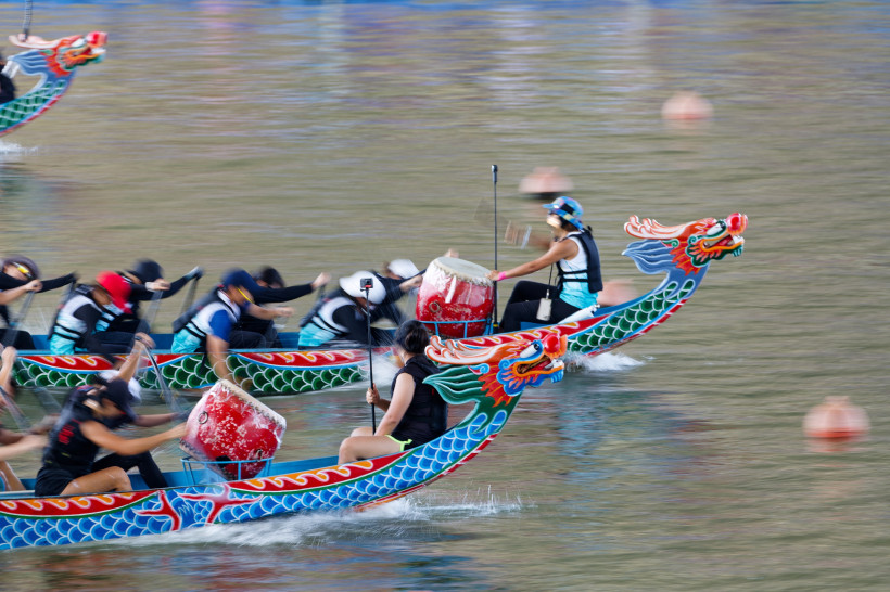 Singapur Drachenboote mit bunten, drachenförmigen Bugfiguren rasen im Wettkampf über das Wasser. Die Paddler bewegen sich synchron, während vorne je ein Trommler den Rhythmus vorgibt. Die Szene wirkt dynamisch und sportlich.