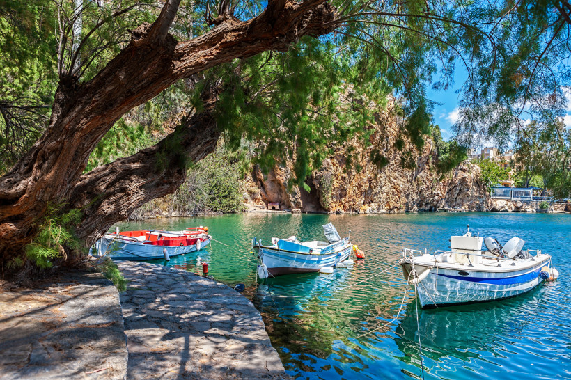 Boote am Ufer des Voulismeni-Sees in Agios Nikolaos, im Vordergrund ein großer Baum und eine Steinpromenade