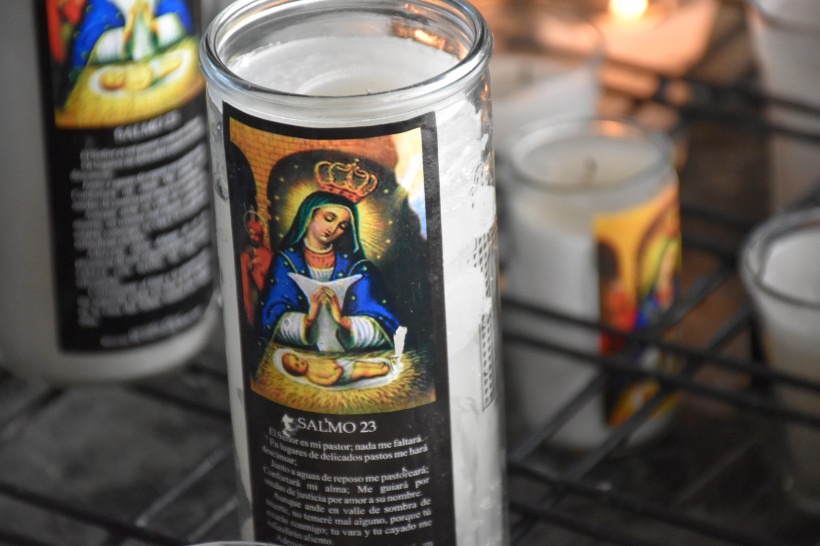 Nahaufnahme einer Votivkerze mit dem Bildnis der Jungfrau Maria mit Krone, die ein Gebet hält, darunter ein schlafendes Jesuskind. Auf der Kerze ist auch Psalm 23 in spanischer Sprache abgedruckt
