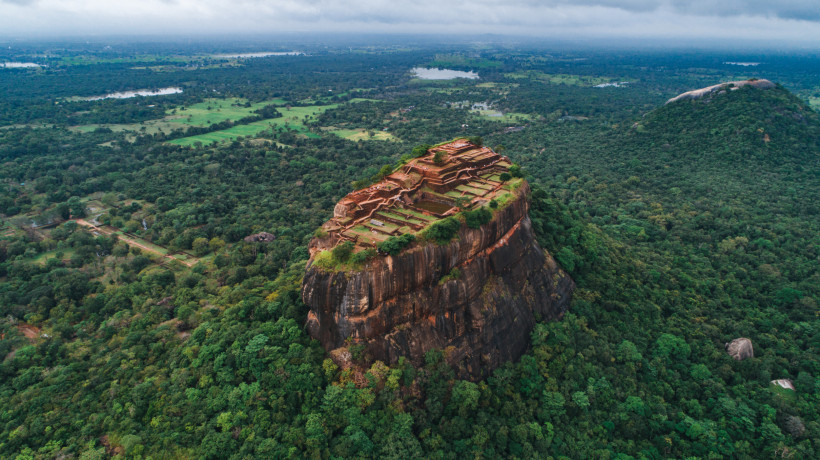 Luftaufnahme der Felsenfestung Sigiriya in Sri Lanka, ein massiver, freistehender Felsen mit gut sichtbaren Ruinen auf dem Gipfel. Die alte Anlage besteht aus Terrassen, Mauern und Gärten. Der Felsen ragt markant aus dem dichten, grünen Dschungel heraus. 