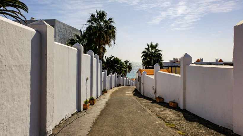 Gasse mit weißen Mauern und Palmen mit Blick auf das Meer an der Playa de Esquinzo