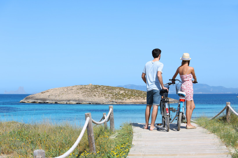 Formentera Ein Paar steht auf einem Holzsteg mit Fahrrädern und blickt auf das türkisblaue Meer und eine kleine Insel in der Ferne. Der Himmel ist klar und blau, und das Paar trägt sommerliche Kleidung
