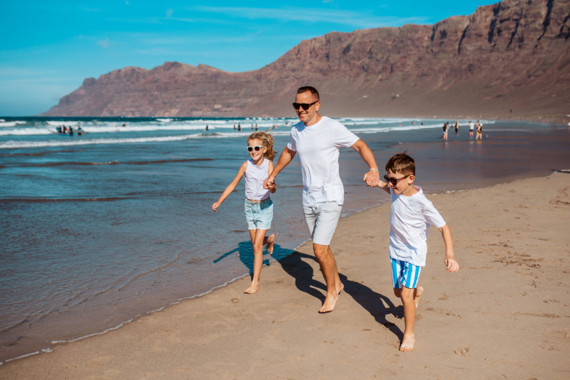 Vater läuft mit zwei Kindern am Strand von Lanzarote und genießt sonniges Familienzeit am Meer.
