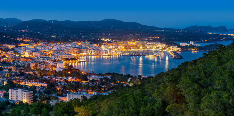 Santa Eulària des Riu Panoramablick auf die Küstenstadt Sant Antoni de Portmany auf Ibiza bei Nacht. Die Lichter der Gebäude spiegeln sich im ruhigen Wasser des Hafens, während bewaldete Hügel im Vordergrund liegen.