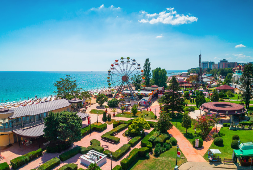 Bulgarien Freizeitpark am Meer mit einem bunten Riesenrad und vielen kleinen Gebäuden. Im Vordergrund gepflegte Grünanlagen, im Hintergrund sieht man zahlreiche Sonnenschirme am Strand und hohe Hotels